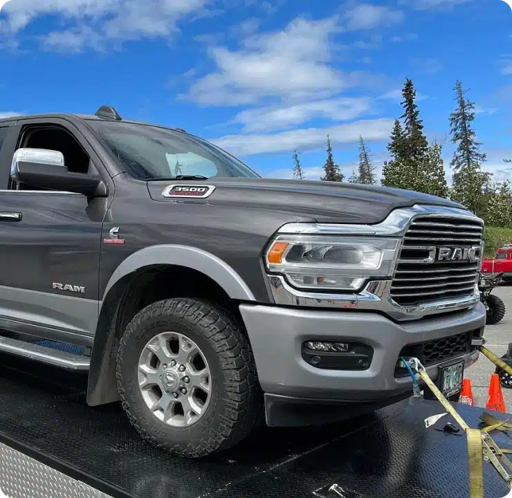 1023 Diesel & Fleet: A gray Ram 2500 truck is parked on a platform, secured with straps. The truck is facing left under a partly cloudy blue sky with trees in the background and orange cones nearby.