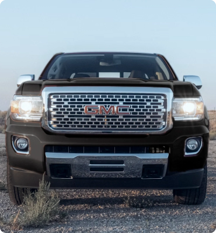 1023 Diesel & Fleet: Front view of a black GMC truck parked on a gravel road with a clear sky background. The large chrome grille and headlights are prominently visible. The foreground features sparse vegetation.