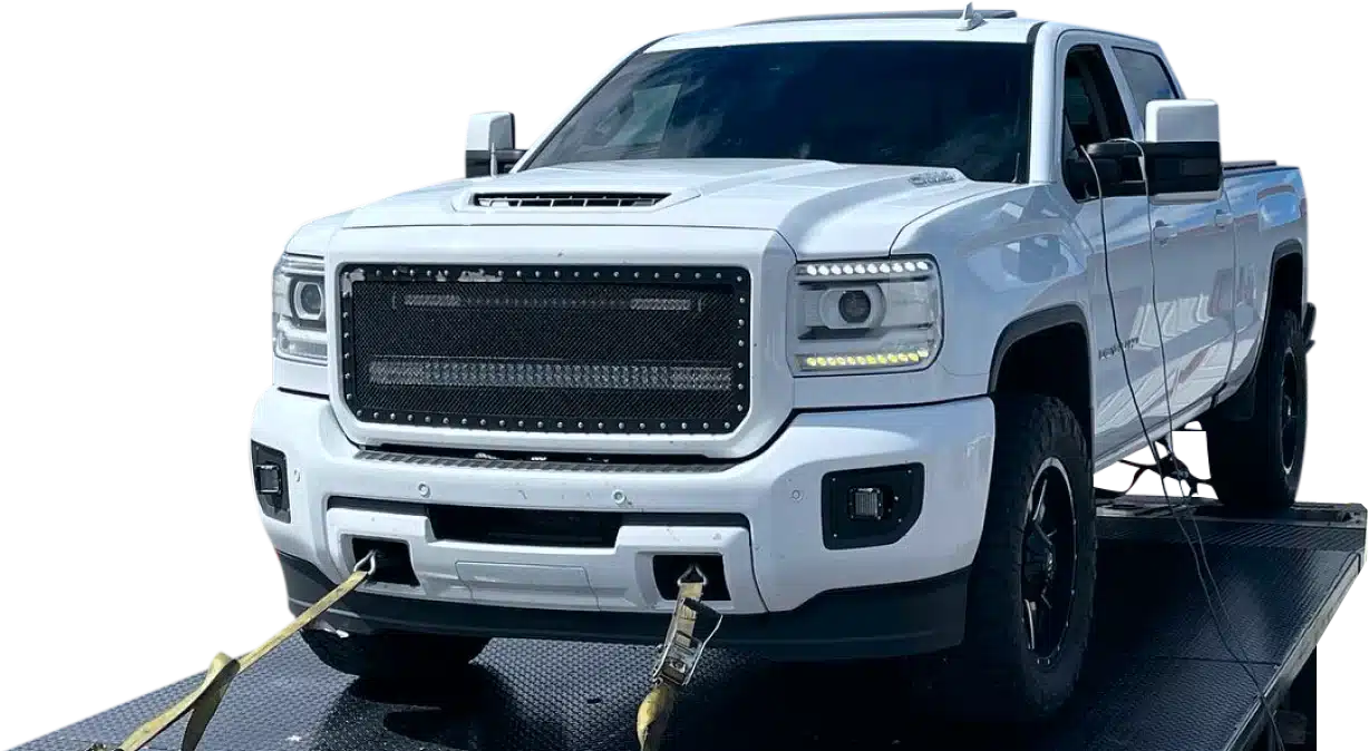 1023 Diesel & Fleet: A white pickup truck with a large front grille is secured on a flatbed trailer using straps. The truck has black wheels and distinctive LED headlights. The background is removed, focusing on the vehicle.