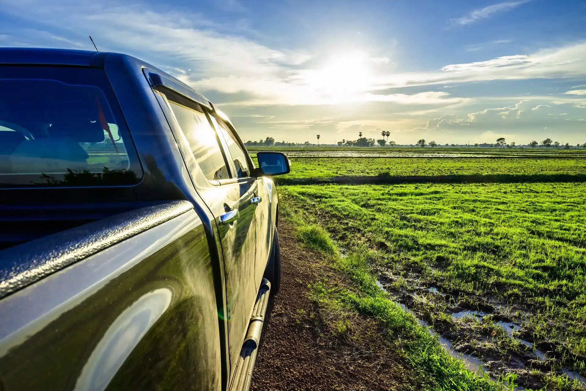 1023 Diesel & Fleet: A pickup truck is parked on a dirt road next to a lush green field under a clear blue sky. The sun is low on the horizon, casting a warm glow over the landscape.