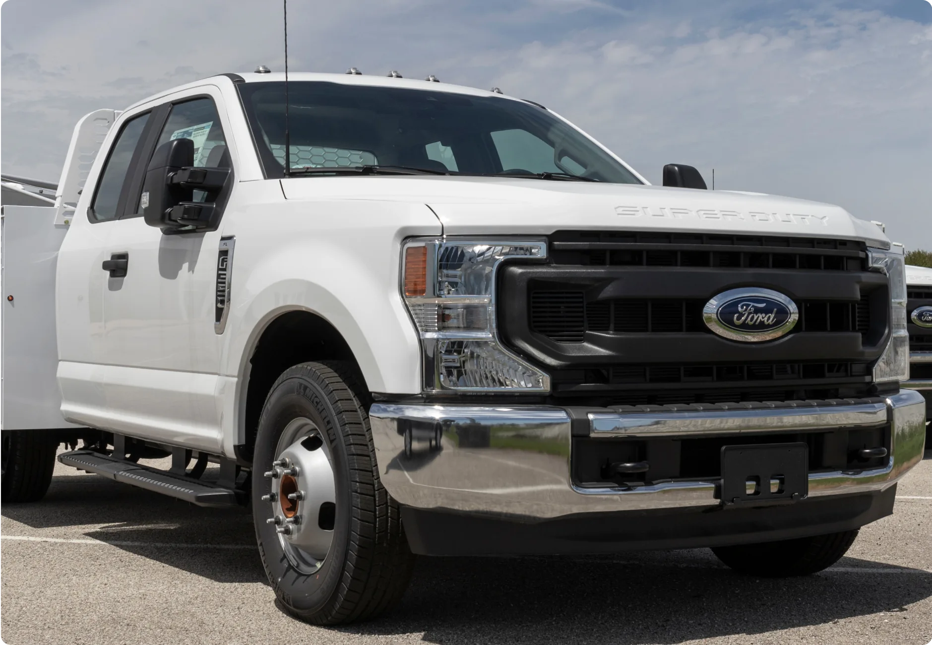 1023 Diesel & Fleet: A white Ford Super Duty truck is parked outdoors on a sunny day. The truck is viewed from a low angle, showcasing its front grille and headlights. The sky in the background is partly cloudy.