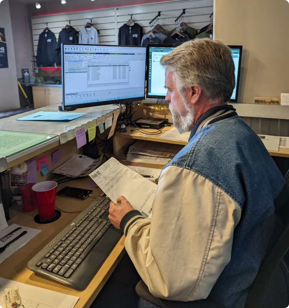 1023 Diesel & Fleet: A man with gray hair and a beard wearing a blue and white jacket sits at a desk with two computer monitors, holding papers. The desk has a keyboard, cup, and notes. Jackets hang in the background.