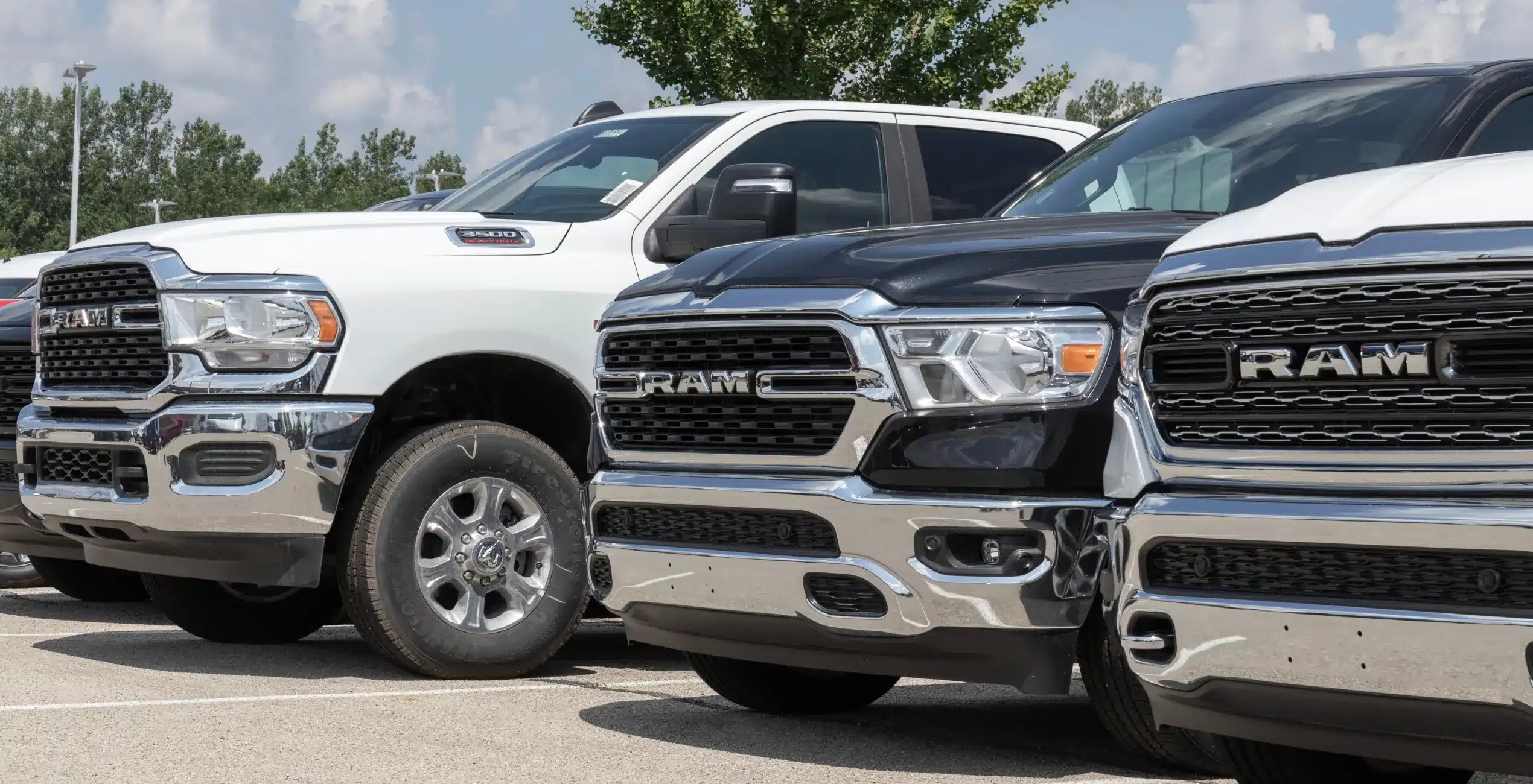 1023 Diesel & Fleet: A row of RAM trucks parked outdoors. The foreground features three trucks in white, black, and gray, with prominent front grilles and chrome accents. Trees and a cloudy sky are visible in the background.