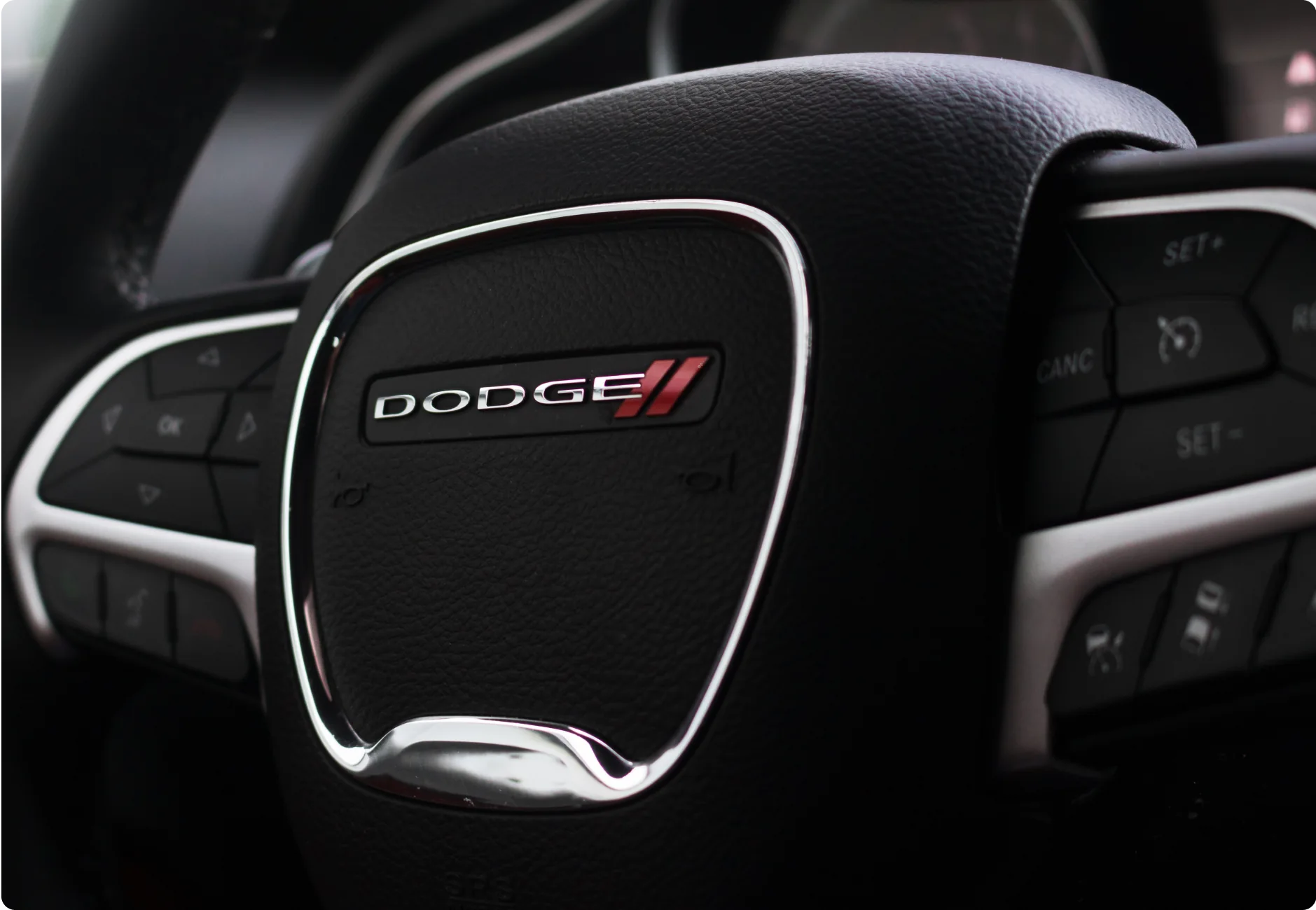 1023 Diesel & Fleet: Close-up of a Dodge car steering wheel, featuring the Dodge logo at the center. The wheel is designed with various control buttons on either side, providing functionality for the driver. The interior appears sleek and modern.