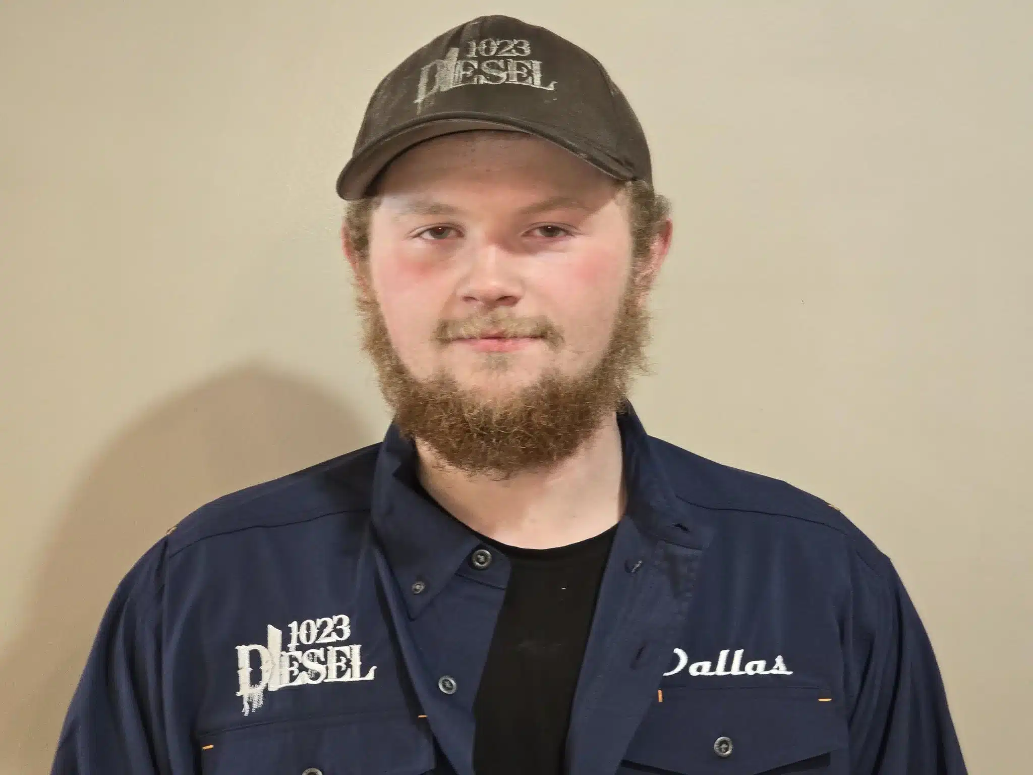 1023 Diesel & Fleet: A bearded individual wearing a black cap with 1023 Diesel written on it, and a blue shirt that has Dallas on the right side, smiles gently against a plain, light-colored background.