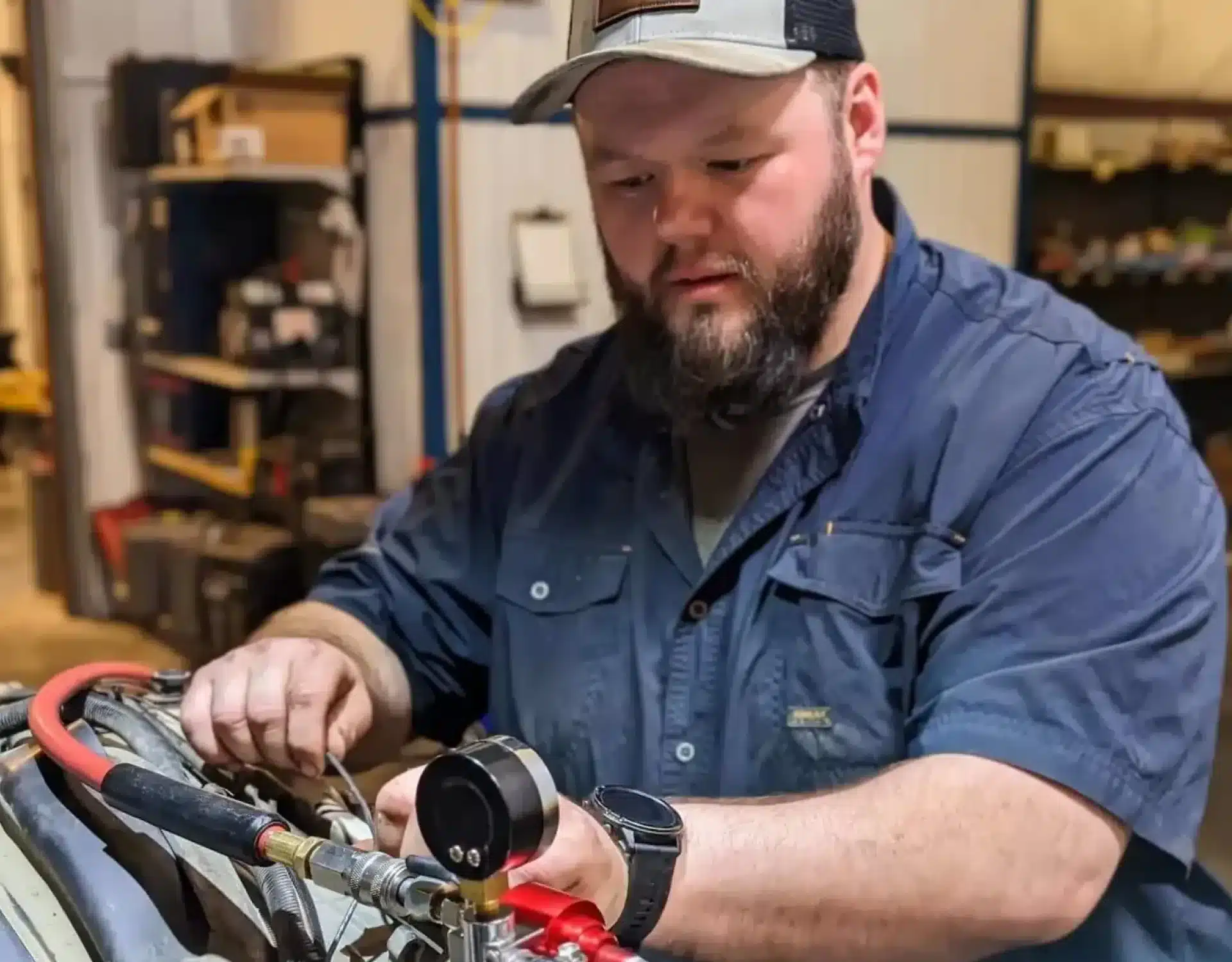 1023 Diesel & Fleet: A man wearing a navy blue shirt and a cap works with tools on machinery in a workshop. He appears focused on adjusting a component, surrounded by various equipment and parts in the background.