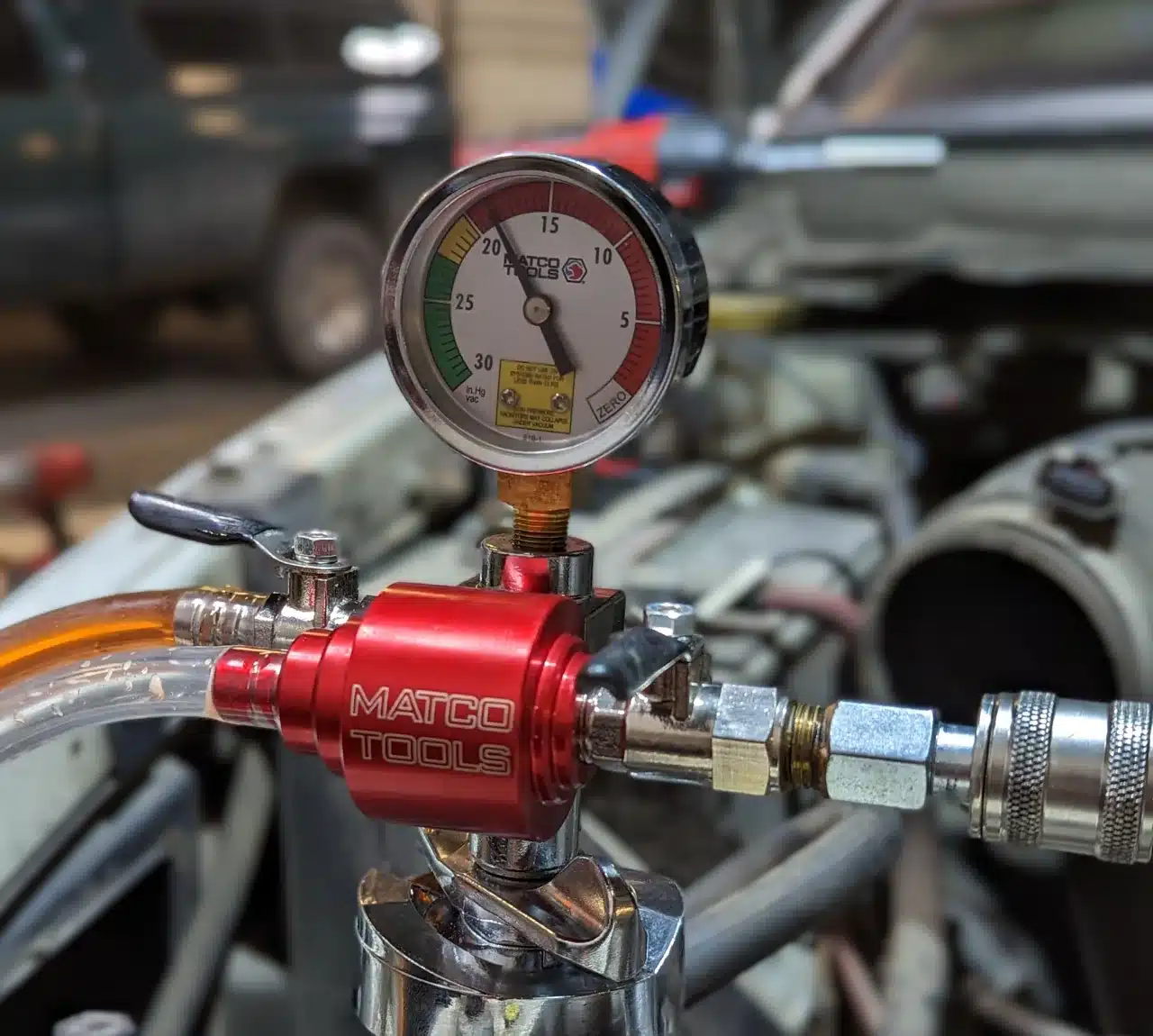 1023 Diesel & Fleet: A pressure gauge from Matco Tools is attached to a red valve, indicating the vacuum level in a car engine. The gauge shows a needle within a colored scale, and the vehicles engine components are visible in the background.