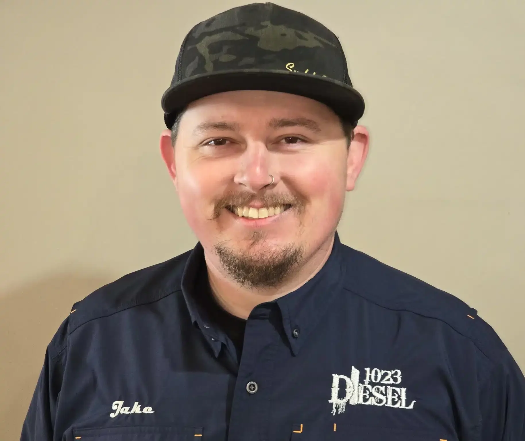 1023 Diesel & Fleet: A person wearing a black cap and a dark shirt that reads Jake and 1023 Diesel smiles at the camera. They have facial hair and a nose ring, standing against a plain background.