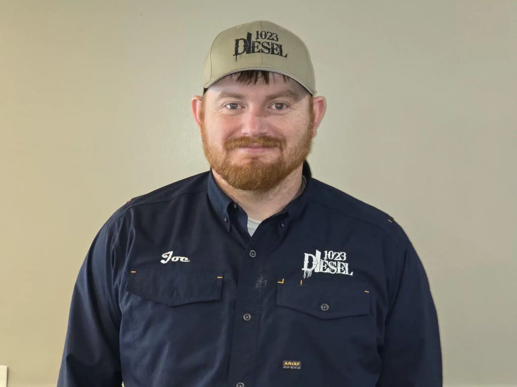 1023 Diesel & Fleet: A man with a beard wearing a light brown cap and a navy blue shirt with 1023 Diesel and Joe embroidered on it stands in front of a gray background.