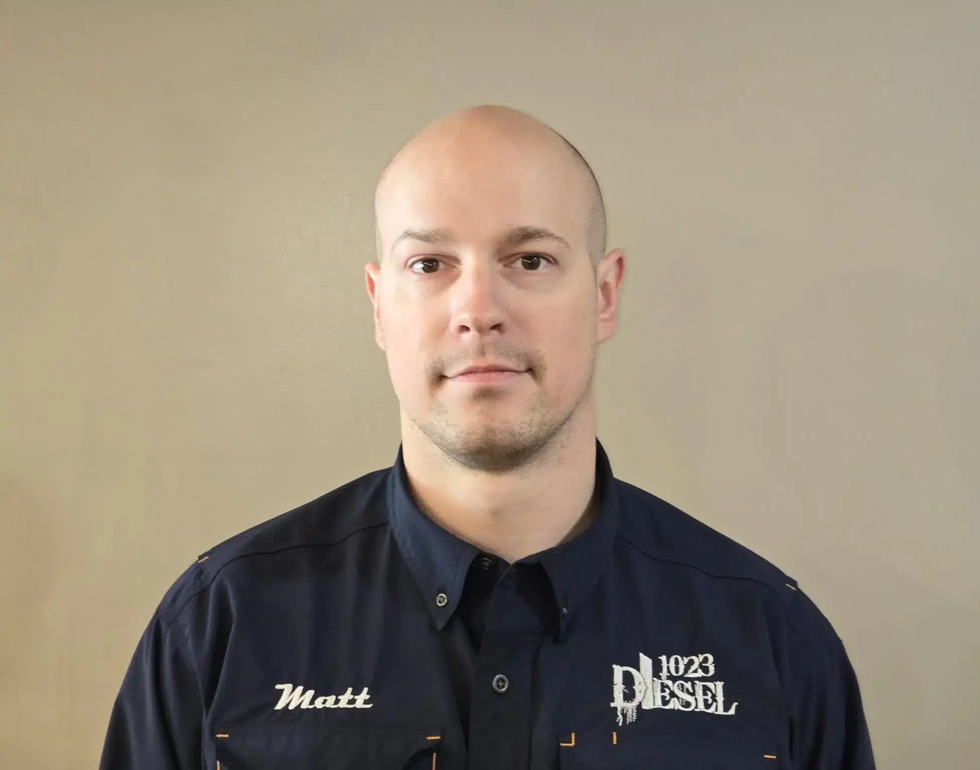 1023 Diesel & Fleet: A man with a shaved head wearing a dark blue shirt with embroidered text Matt and 1023 Diesel stands in front of a plain beige background.