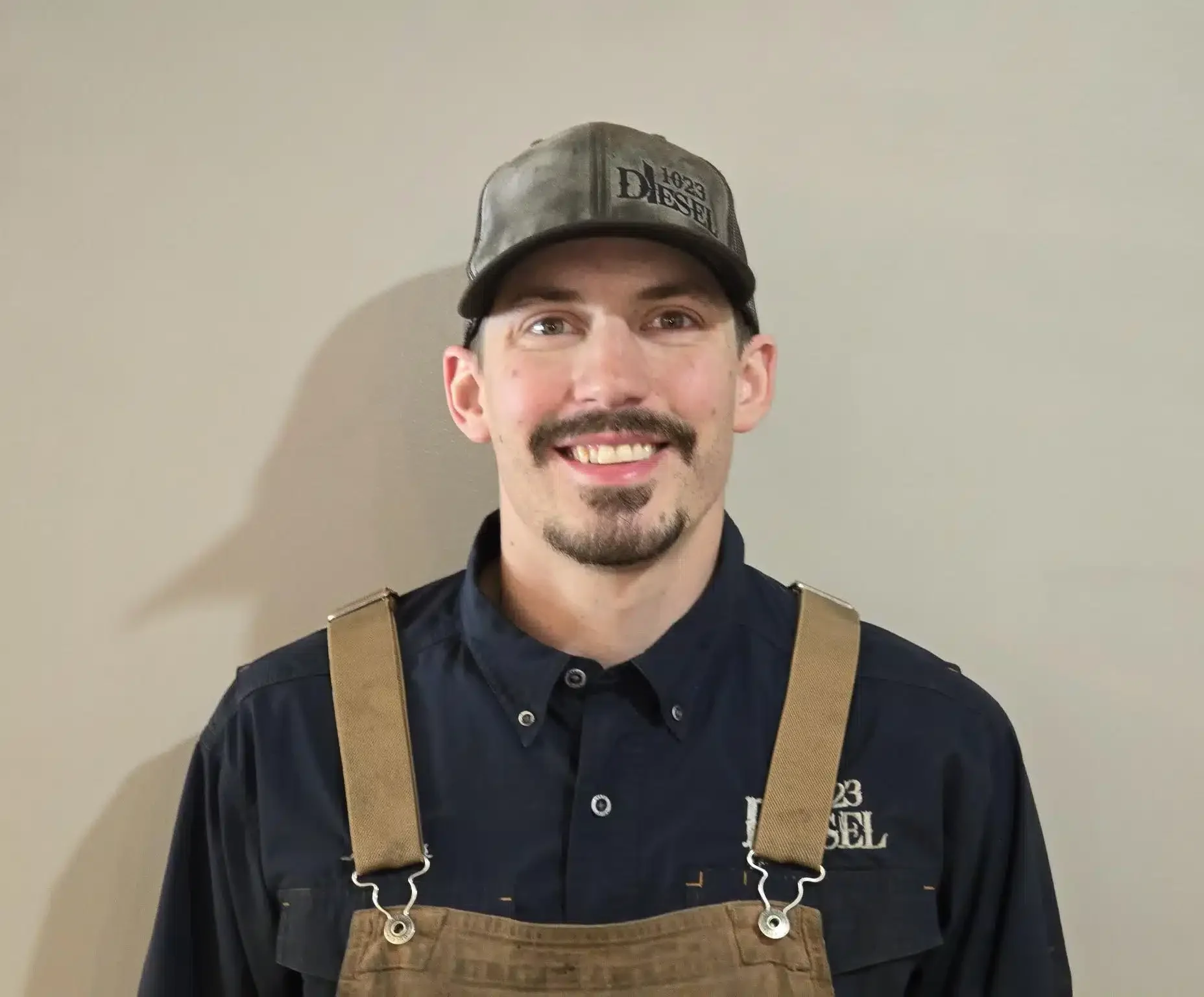 1023 Diesel & Fleet: A man wearing a black cap, navy shirt, and brown suspenders smiles against a plain background. His shirt and cap feature the same brand name.