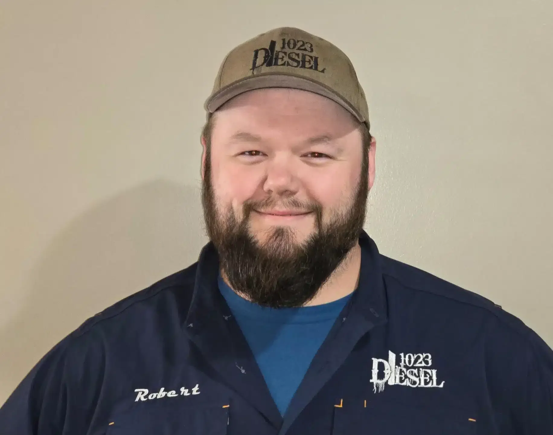 1023 Diesel & Fleet: Man with a beard wearing a brown cap and a blue shirt with embroidered text Robert and 1023 Diesel. He is standing against a plain background, smiling softly at the camera.