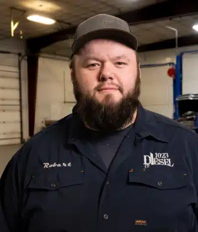 carrers, diesel repair in Palmer, AK at 1023 Diesel & Fleet: A man with a beard wearing a cap and a dark shirt with 1023 Diesel and a name tag on it stands in a garage with a partially visible vehicle in the background. The lighting is from overhead lights.