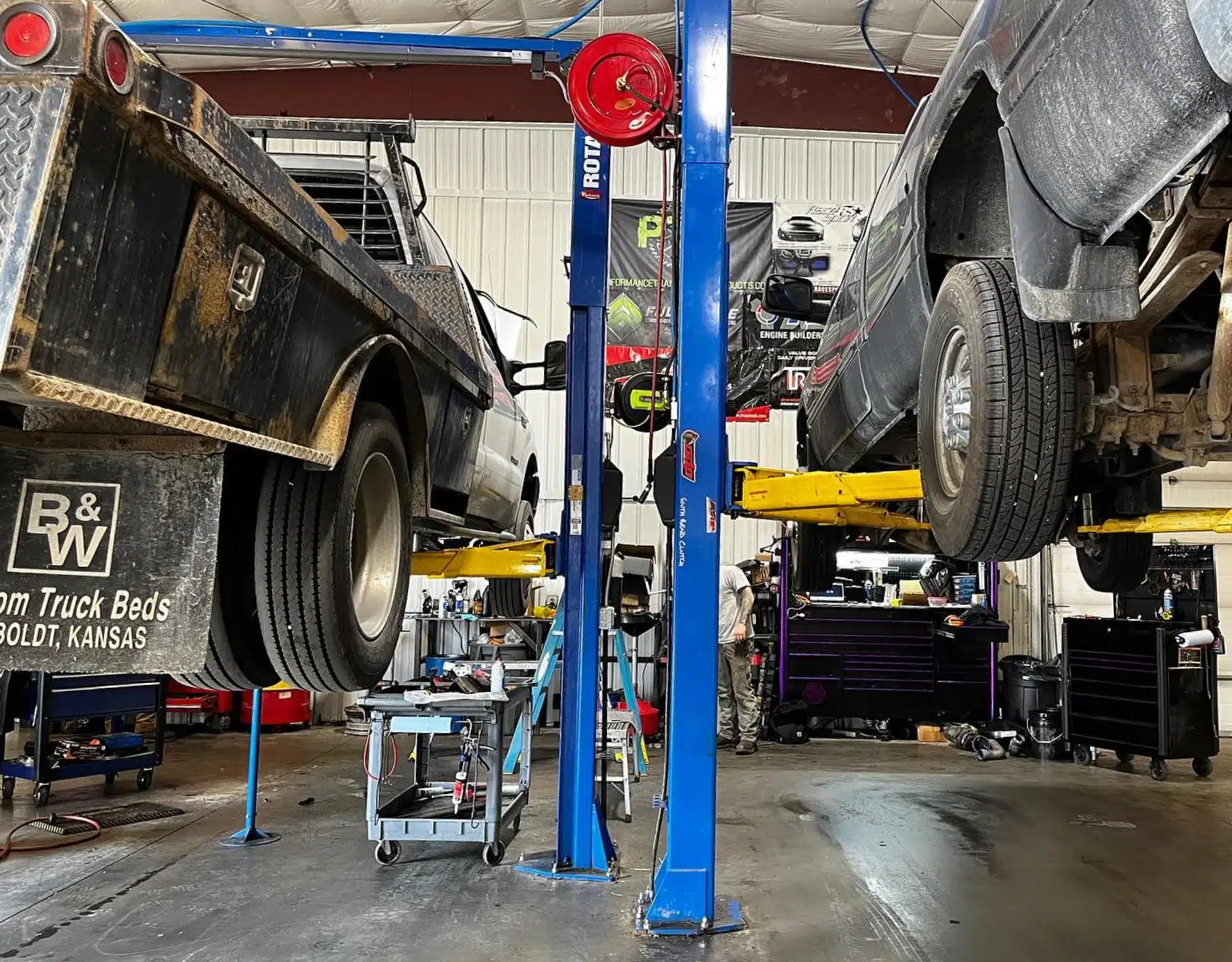 1023 Diesel & Fleet: Two trucks are lifted on hydraulic lifts in a garage. The trucks are visibly dirty, with one having a B&W Custom Truck Beds sign. The garage has tools and equipment, with a worker visible in the background.