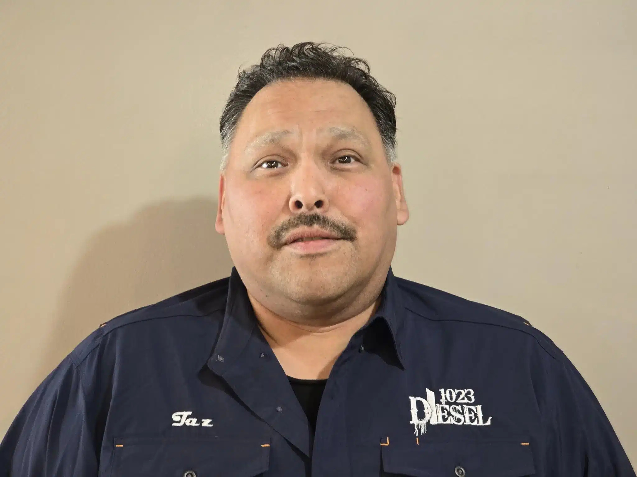 1023 Diesel & Fleet: A man with short dark hair and a mustache is wearing a dark blue shirt with embroidered text that reads Taz and 1023 Diesel. The background is a neutral beige color.