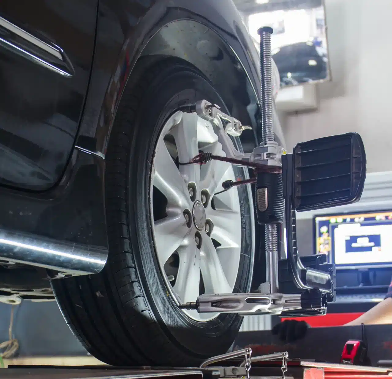 1023 Diesel & Fleet: Close-up of a black car undergoing wheel alignment in a garage. A device is attached to the wheel, and a computer screen is visible in the background, displaying alignment details.