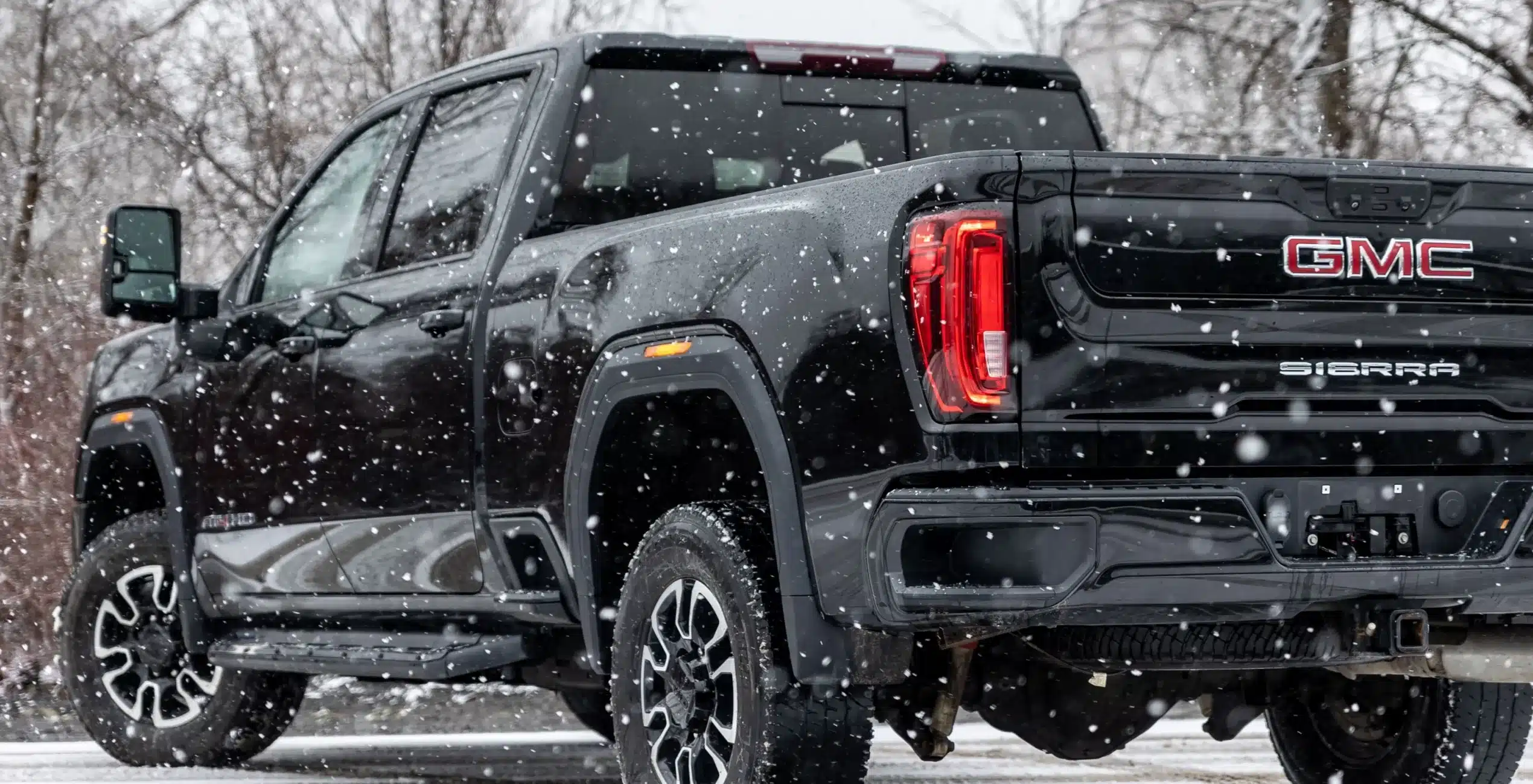 1023 Diesel & Fleet: A black GMC Sierra truck is pictured from the rear on a snowy day. Snowflakes are falling, and trees with bare branches are visible in the background. The trucks taillights are illuminated against the wintry scene.