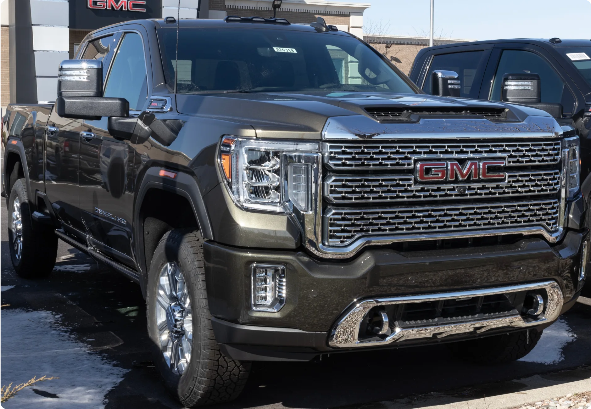 1023 Diesel & Fleet: A black GMC Sierra 2500HD pickup truck is parked in a lot. The truck features a large grille with chrome accents, LED headlights, and a muscular hood. Snow is visible on the ground, and another vehicle is parked next to it.