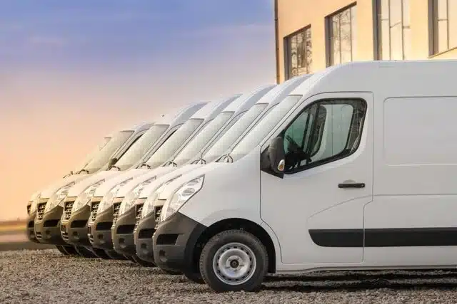 1023 Diesel & Fleet: A row of identical white delivery vans parked on gravel beside a building. The sky is clear with a warm sunset or sunrise hue.