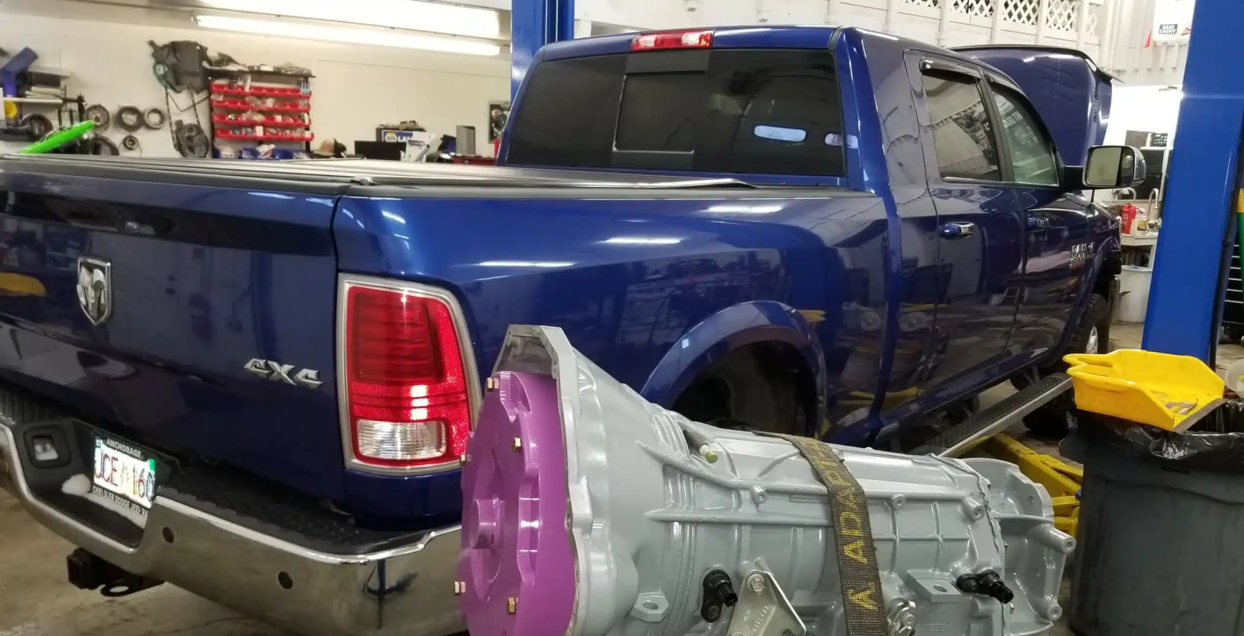 1023 Diesel & Fleet: A blue pickup truck is parked in a garage workshop with its hood open. In the foreground, a transmission with a purple component is visible, resting on the ground. Various tools are scattered around the workshop.