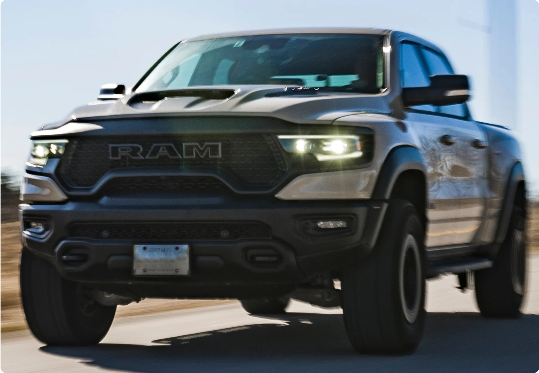 1023 Diesel & Fleet: A silver RAM pickup truck driving on a road, with headlights on and motion blur emphasizing speed. The vehicle has a prominent front grille and large tires. The background is blurred, suggesting movement.