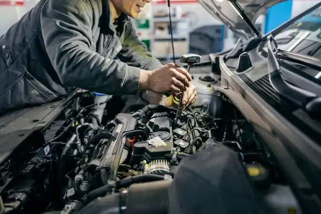 1023 Diesel & Fleet: A mechanic in a gray jumpsuit works under the hood of a car, using a wrench on the engine. The workshop setting includes various tools and equipment in the background, indicating a repair or maintenance task in progress.