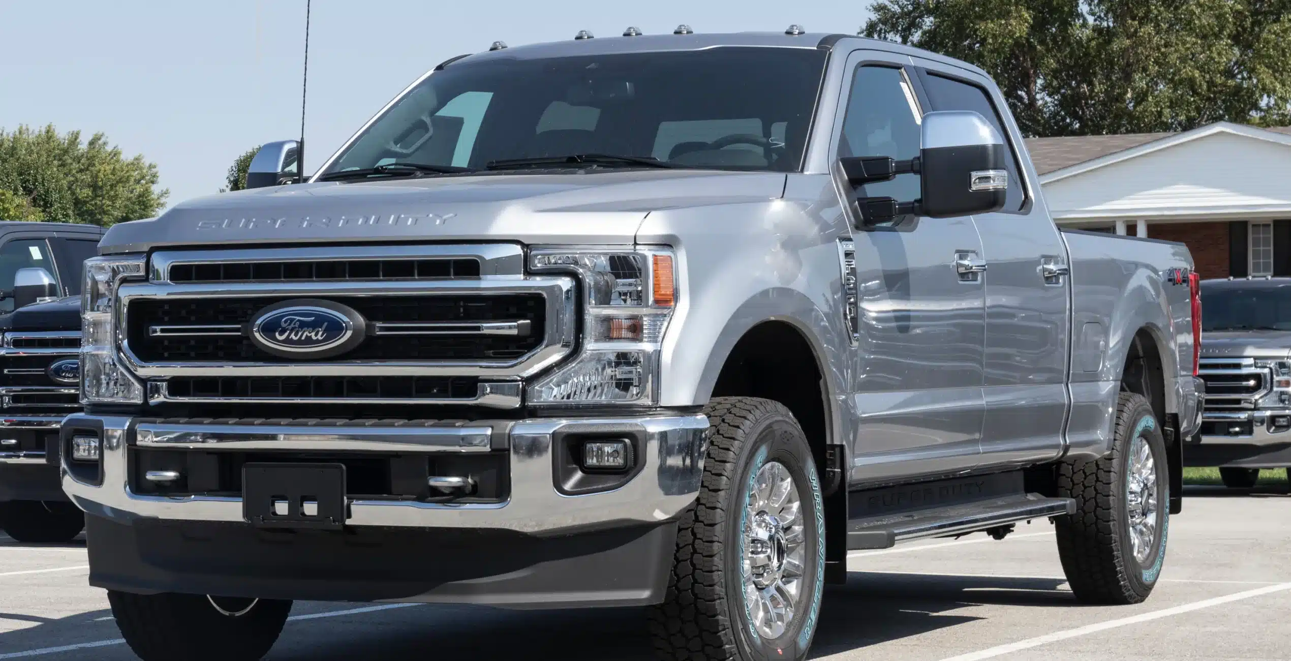 1023 Diesel & Fleet: A silver Ford Super Duty pickup truck is parked on a paved surface near a residential area. The trucks bold grille and large side mirrors are prominent, and other vehicles are visible in the background.