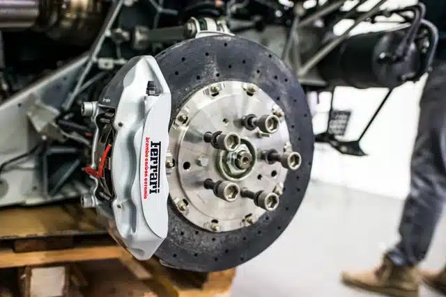 1023 Diesel & Fleet: Close-up of a cars brake system, featuring a vented disc and a white caliper with red Ferrari logo. The background shows part of a vehicles suspension system and a person slightly out of focus.