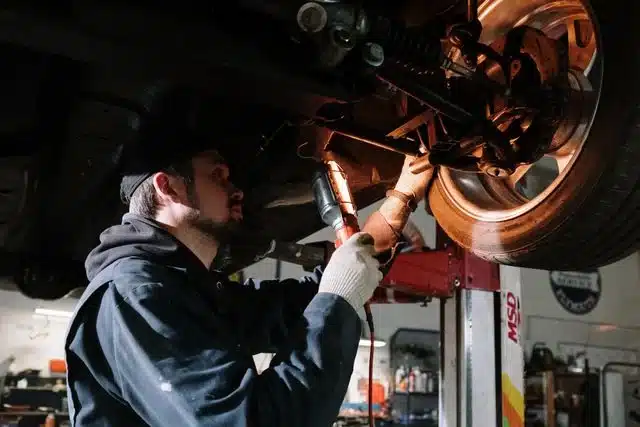 1023 Diesel & Fleet: A mechanic working under a car on a lift, focusing on the wheel area. He is wearing a cap, gloves, and protective jacket, and is using a tool in a well-lit garage with various equipment in the background.