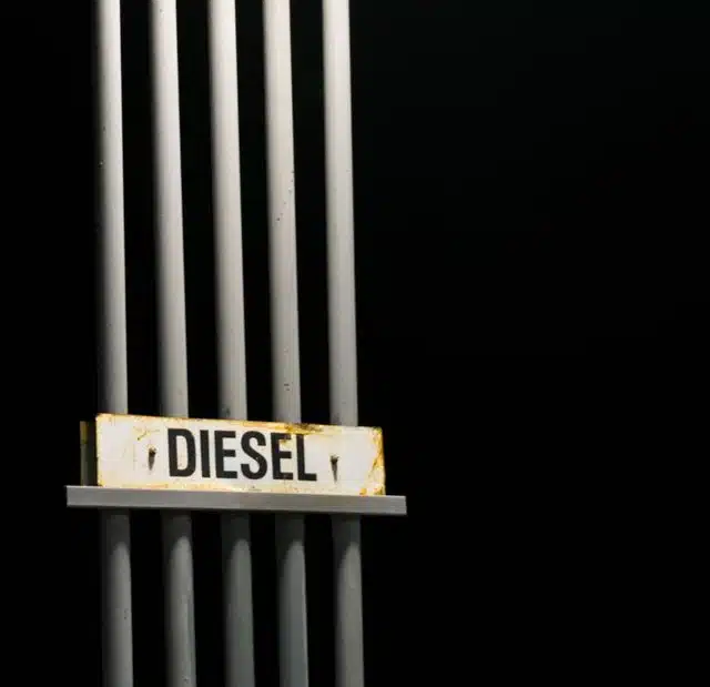 1023 Diesel & Fleet: A close-up of a weathered sign with the word DIESEL attached to a vertical structure of metal pipes against a dark background. The pipes are evenly spaced and cast soft shadows, highlighting the industrial feel of the image.