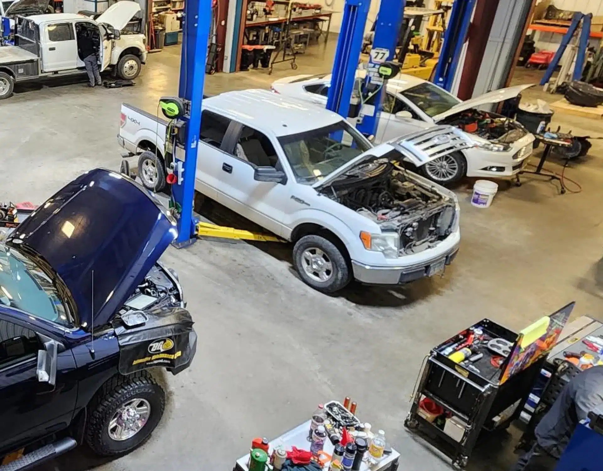 1023 Diesel & Fleet: A busy auto repair shop with several vehicles on hydraulic lifts. Mechanics work on different cars, with hoods open and tools scattered around. The scene includes a white truck, a dark truck, and a silver car, along with various equipment.