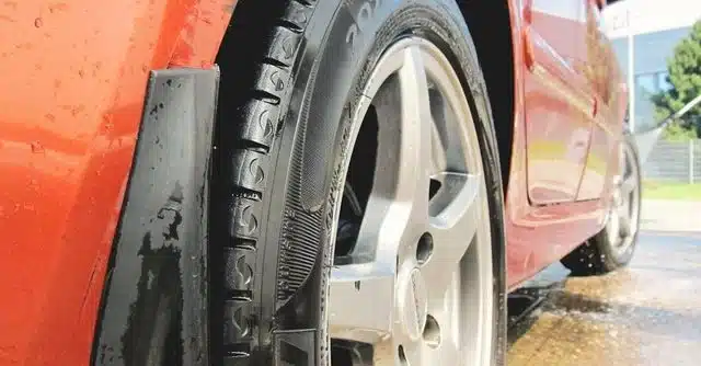 1023 Diesel & Fleet: Close-up view of a red car wheel with shiny metal rims, wet from water. The background shows a blurred outdoor scene with greenery and a building, suggesting the car is being washed.