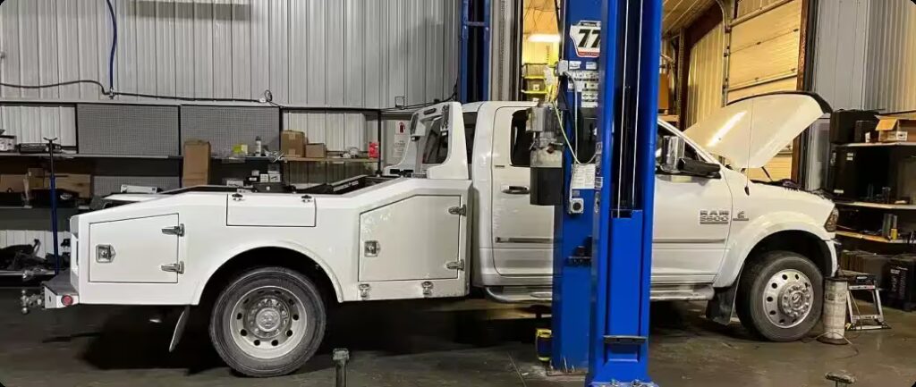 1023 Diesel & Fleet: A white Ram 3500 truck is on a lift in a garage with its hood open. The garage has tools, shelves, and equipment scattered around. The truck appears to be undergoing maintenance or repair work.