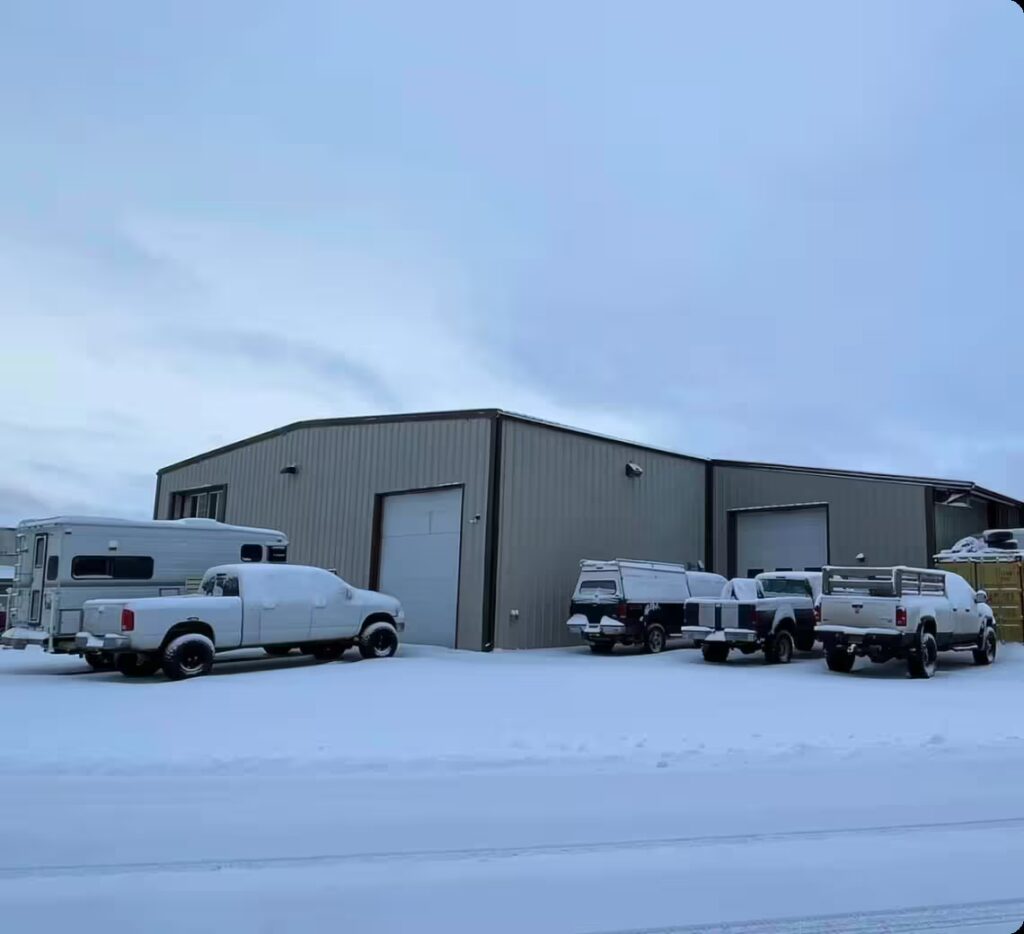 1023 Diesel & Fleet: A large metal warehouse is seen in a snowy landscape. Several vehicles, including vans and trucks, are parked in front, covered with snow. The sky is overcast, creating a muted, wintry atmosphere.