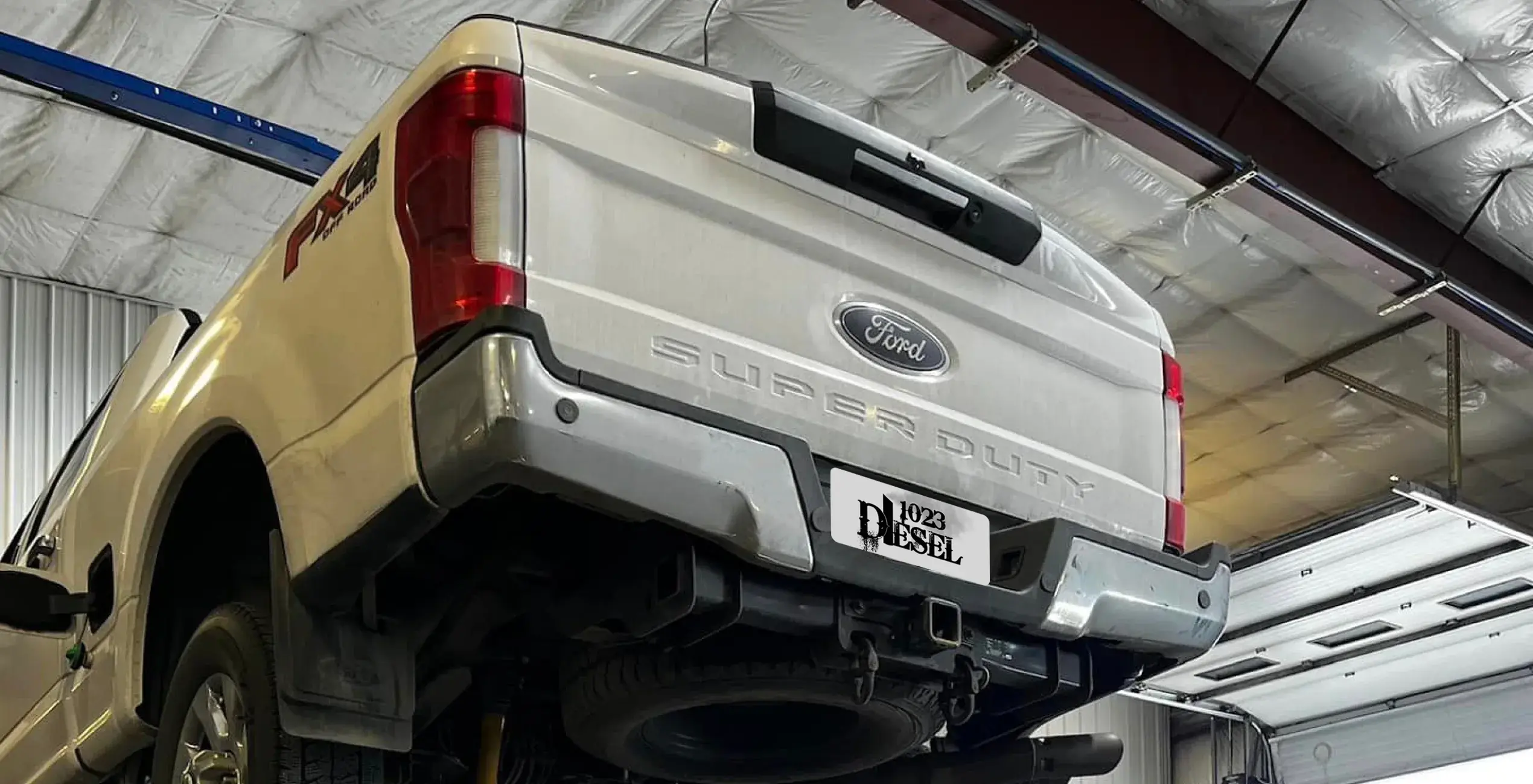 1023 Diesel & Fleet: A white Ford Super Duty truck is lifted in a garage. The rear of the truck is visible, showing the tailgate, bumper, and hitch. The ceiling of the garage features exposed beams and insulation.