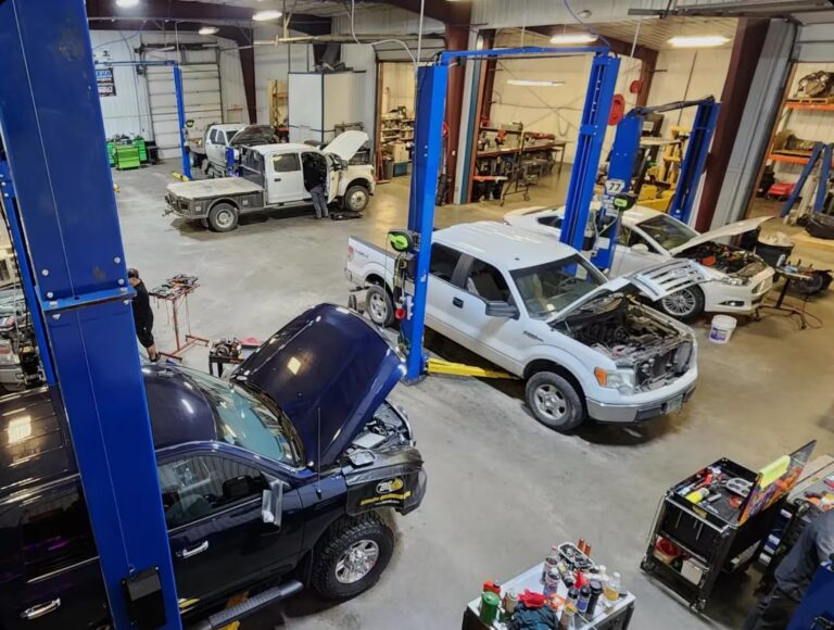 1023 Diesel & Fleet: A busy auto repair shop with multiple vehicles being serviced. Several mechanics work on cars with open hoods, surrounded by tools and equipment. Car lifts are visible, and the workshop is well-lit and organized.