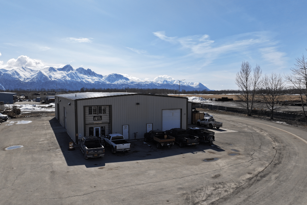 diesel shops, diesel repair in Palmer, AK at 1023 Diesel & Fleet. Wide-angle view of an auto repair shop building with trucks parked outside and snow-capped mountains in the background.