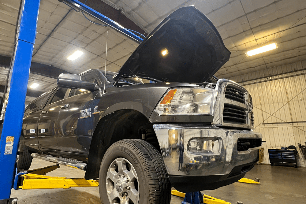 Diesel Maintenance in Palmer, AK at 1023 Diesel & Fleet. A large black pickup truck is lifted on a hydraulic car lift inside an automotive repair shop. The truck’s hood is open, and the garage is brightly lit with tools and equipment visible in the background