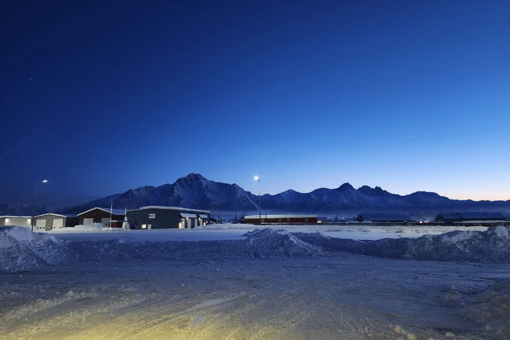 Diesel starting problems in Palmer, AK at 1023 Diesel & Fleet. Image of a snow-covered industrial area with buildings and streetlights in the foreground, framed by rugged mountain peaks under a deep blue twilight sky, conveying cold, quiet winter conditions.