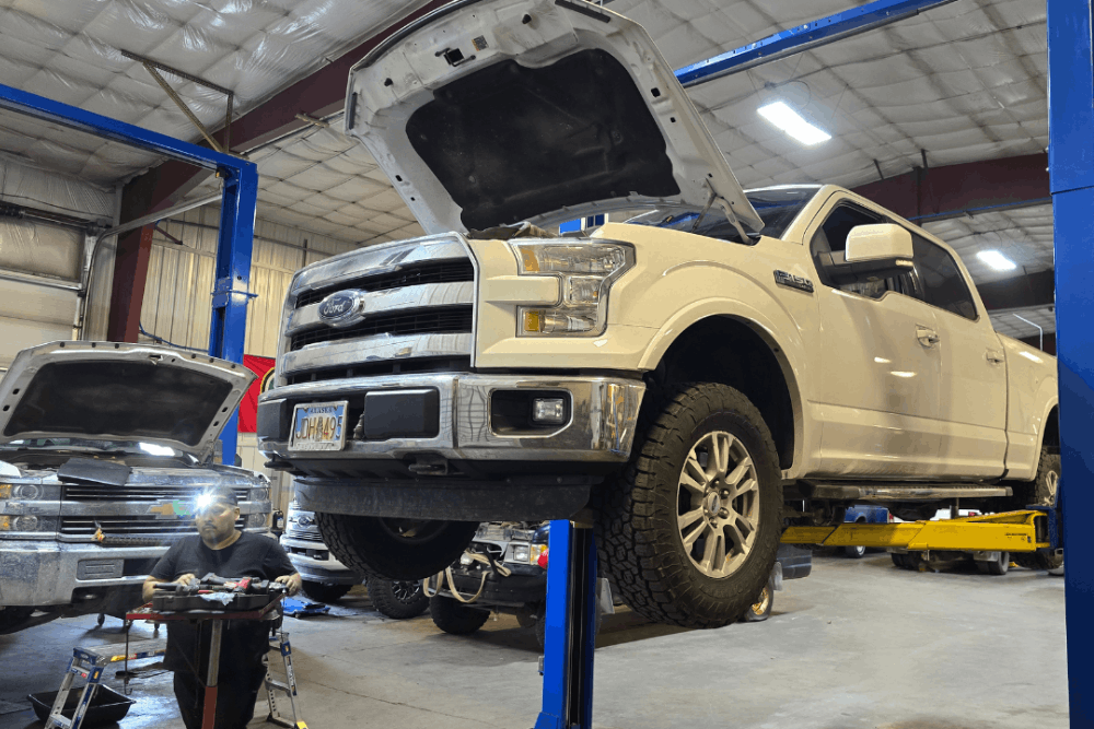 Towing Heavy in Alaska? Prevent Diesel Transmission & Cooling Failures - 1023 Diesel and Fleet - White pickup truck on a blue hydraulic lift in a large garage, hood open, with a mechanic working nearby. Brightly lit with a busy workshop vibe.