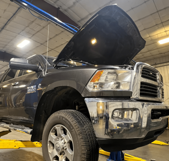 Diesel Maintenance in Palmer, AK at 1023 Diesel & Fleet. A large black pickup truck is lifted on a hydraulic car lift inside an automotive repair shop. The truck’s hood is open, and the garage is brightly lit with tools and equipment visible in the background