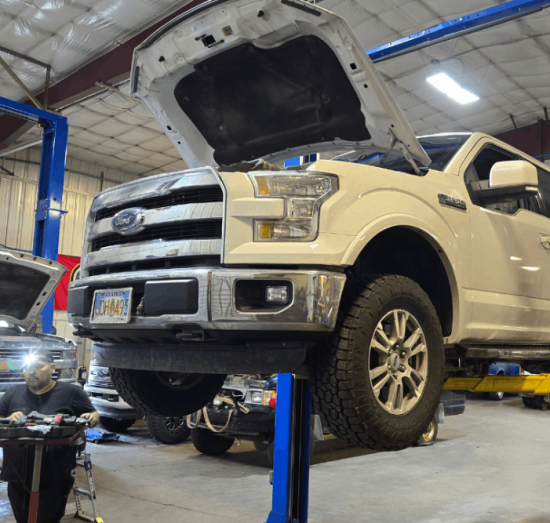 Towing Heavy in Alaska? Prevent Diesel Transmission & Cooling Failures - 1023 Diesel and Fleet - White pickup truck on a blue hydraulic lift in a large garage, hood open, with a mechanic working nearby. Brightly lit with a busy workshop vibe.