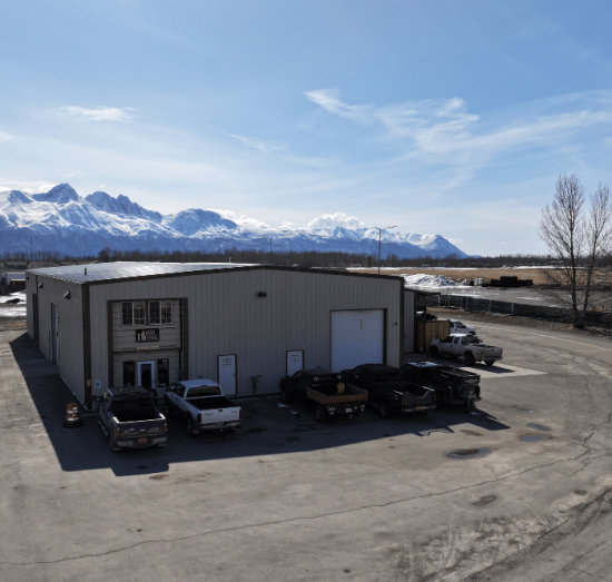 diesel shops, diesel repair in Palmer, AK at 1023 Diesel & Fleet. Wide-angle view of an auto repair shop building with trucks parked outside and snow-capped mountains in the background.