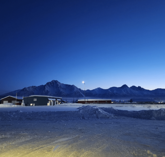 Diesel starting problems in Palmer, AK at 1023 Diesel & Fleet. Image of a snow-covered industrial area with buildings and streetlights in the foreground, framed by rugged mountain peaks under a deep blue twilight sky, conveying cold, quiet winter conditions.
